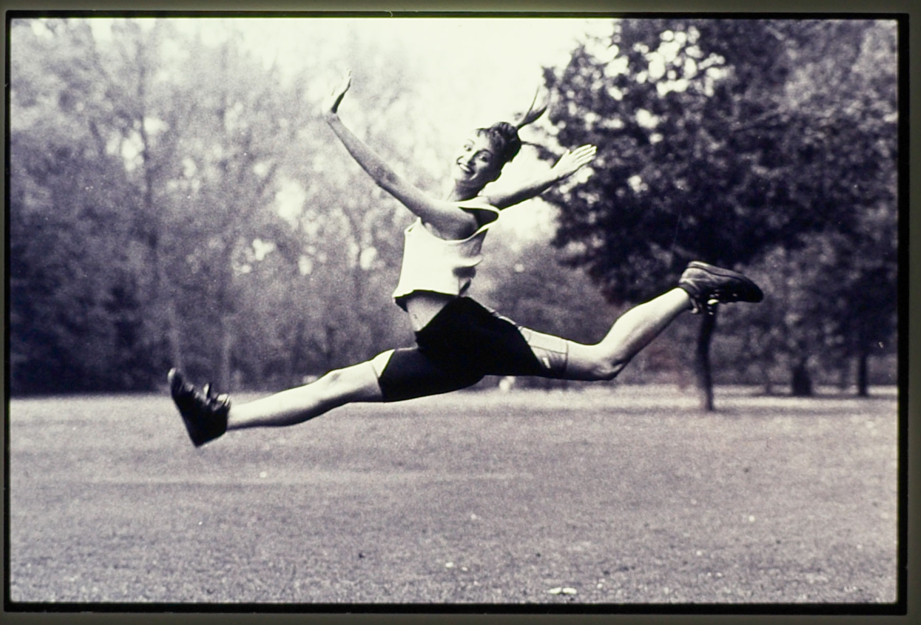 Eine Frau springt mit gestreckten Beinen über eine Wiese. Sie trägt Sportkleidung. Ein Mann mit Kontrabass. Er blickt nach unten. Das Bild ist analog in schwarzweiss aufgenommen. Fotograf: Jörg Saibou, Köln Ehrenfeld